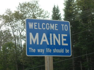 Blue rectangular sign on a wooden post against a background of deciduous and confier trees. White lettering on the sign reads "Welcome to Maine: The Way Life Should Be."